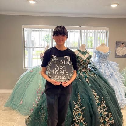 young woman holding a sign that says I said Yes to the Dress at a formal wear shop standing infront of several ball gowns