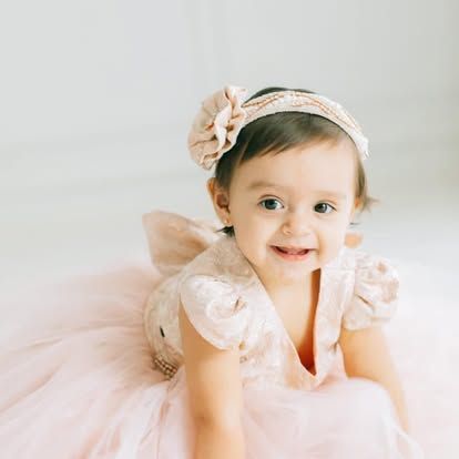 A baby girl in a pink dress and headband is crawling on the floor.