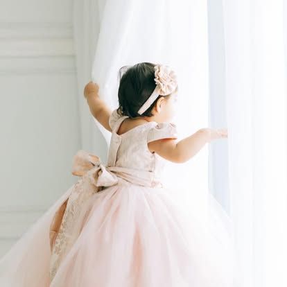 A little girl in a pink dress is standing in front of a window.