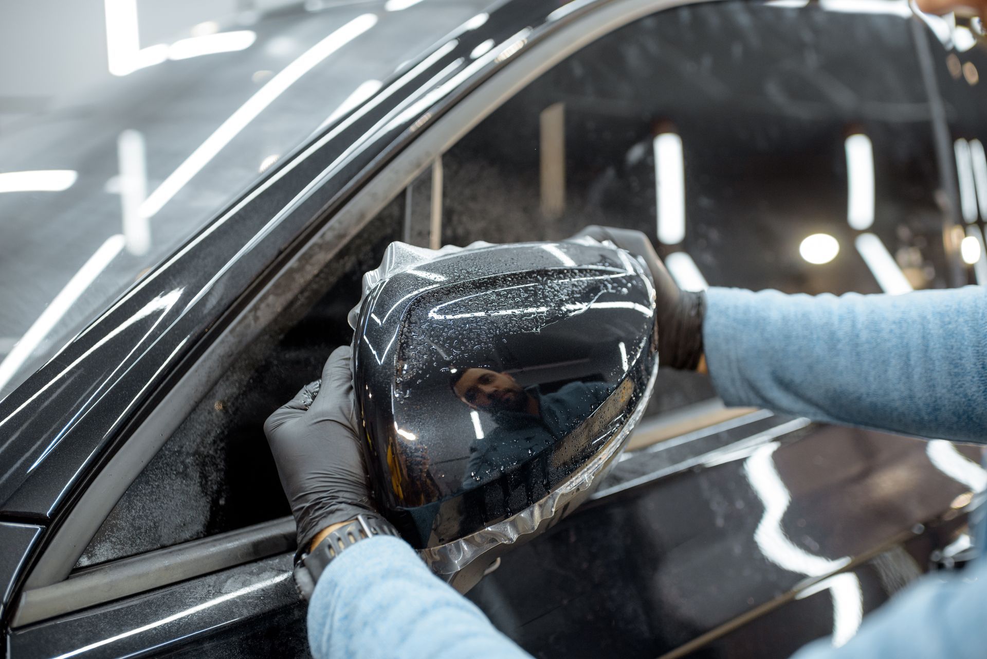 Person in black gloves applying film to a black car mirror indoors.