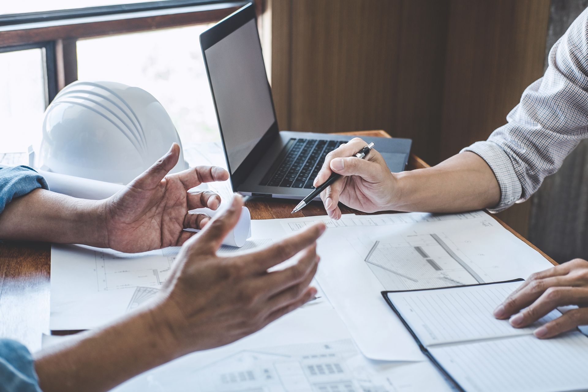 Two people discussing architectural plans at a desk with a laptop and hard hat.