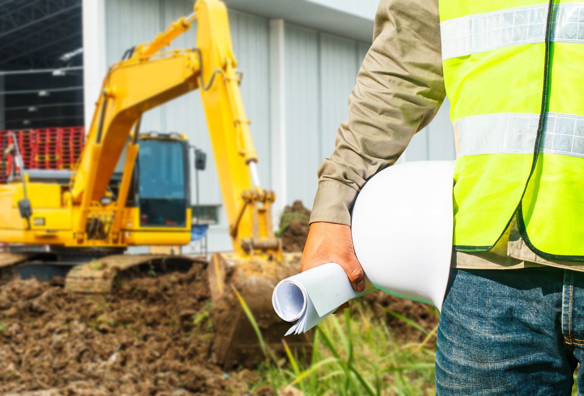 Construction worker holding blueprints, standing near a yellow excavator at a construction site.
