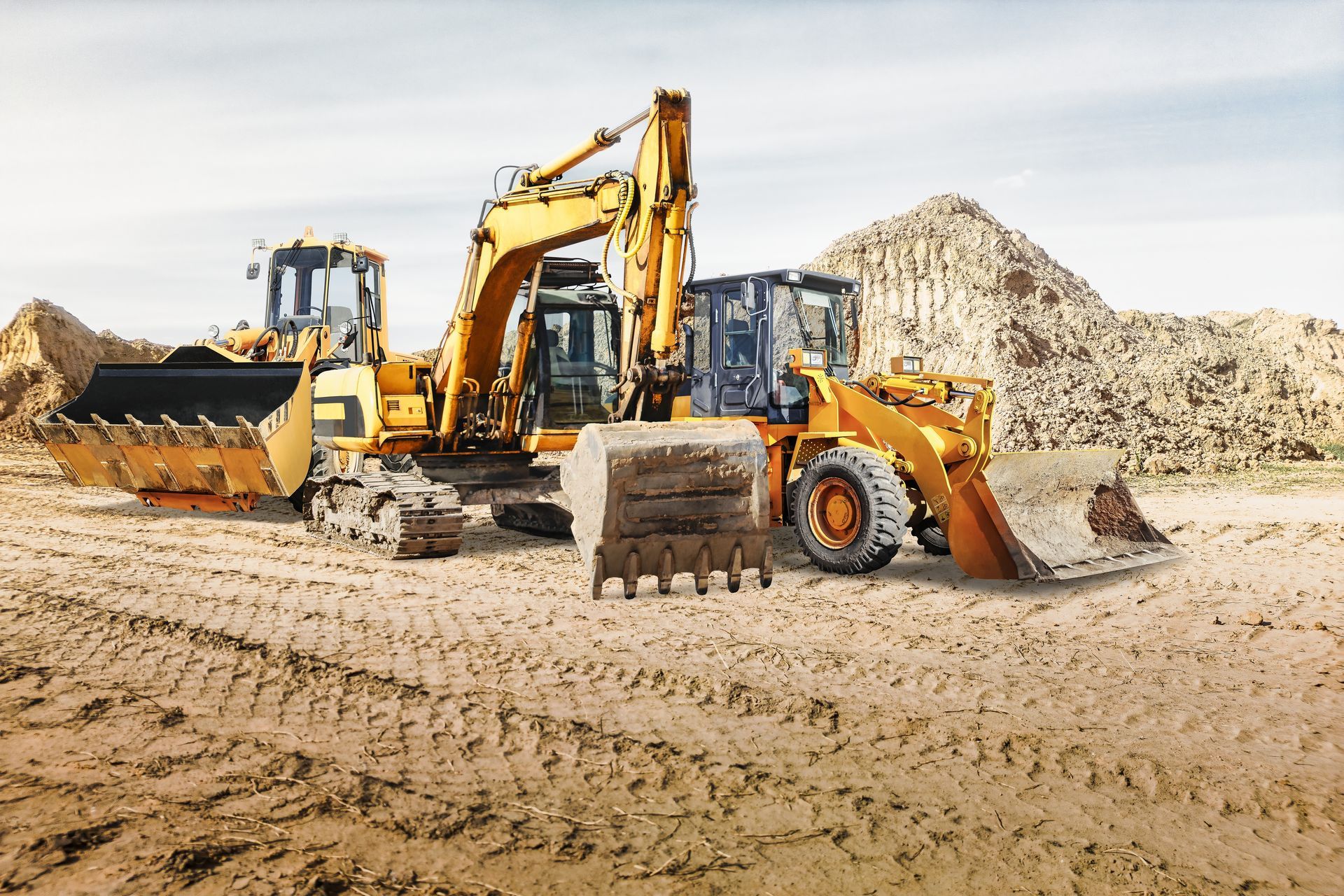Yellow construction vehicles on a dirt construction site, including a backhoe and loader near dirt piles.