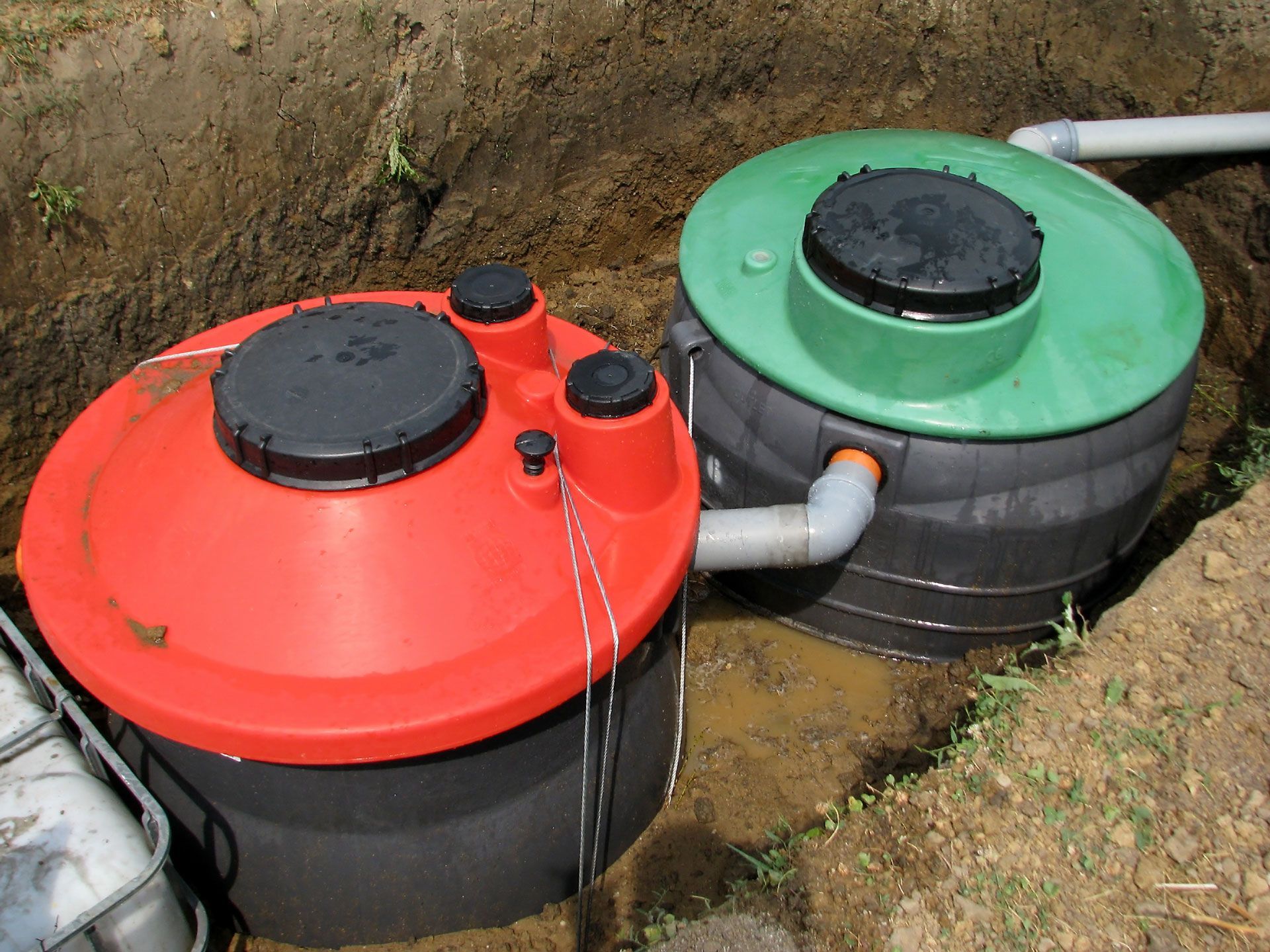 Two round plastic sewage tanks, one red and one green, are installed side-by-side in a dirt trench, connected by a pipe.