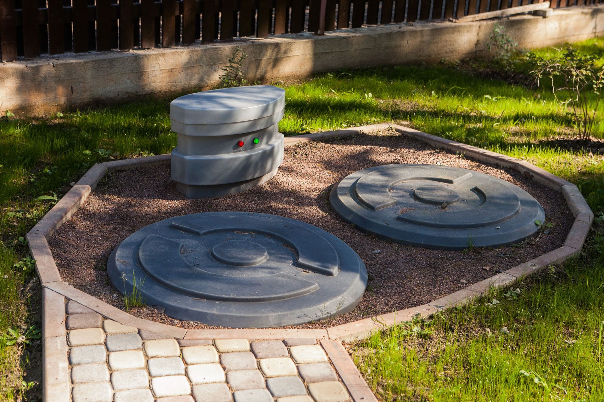 Two gray septic tank access covers and a small control box located in a landscaped backyard setting.