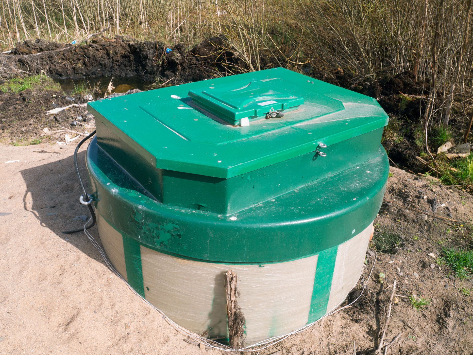 A green-lidded, circular concrete sewage treatment tank or septic system set in a sandy, outdoor landscape.