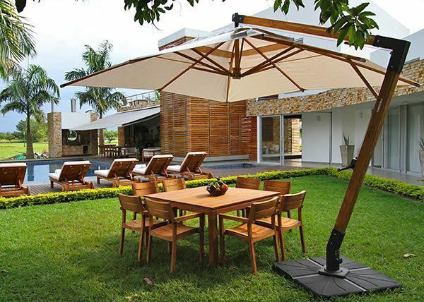 Patio con mesa, sillas y una gran sombrilla. Césped verde, piscina y casa al fondo.