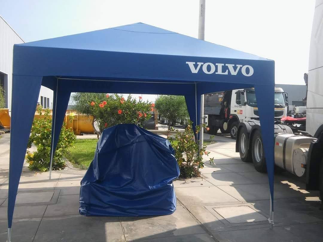 Toldo azul de la marca Volvo con artículo cubierto con una lona, ​​cerca de un camión y arbustos, al aire libre.