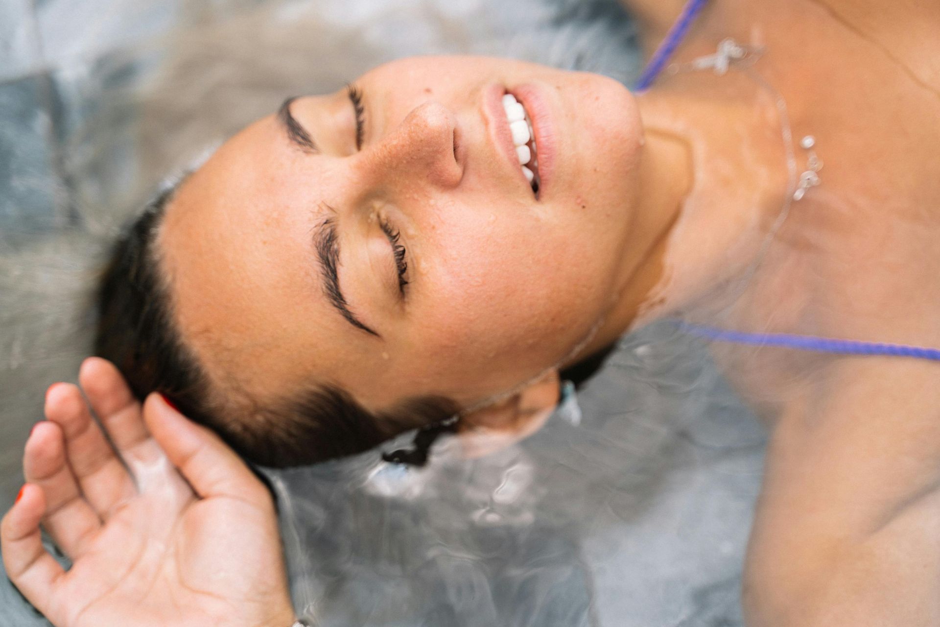 Woman relaxing in water, eyes closed, hand outstretched, wearing a purple strap.