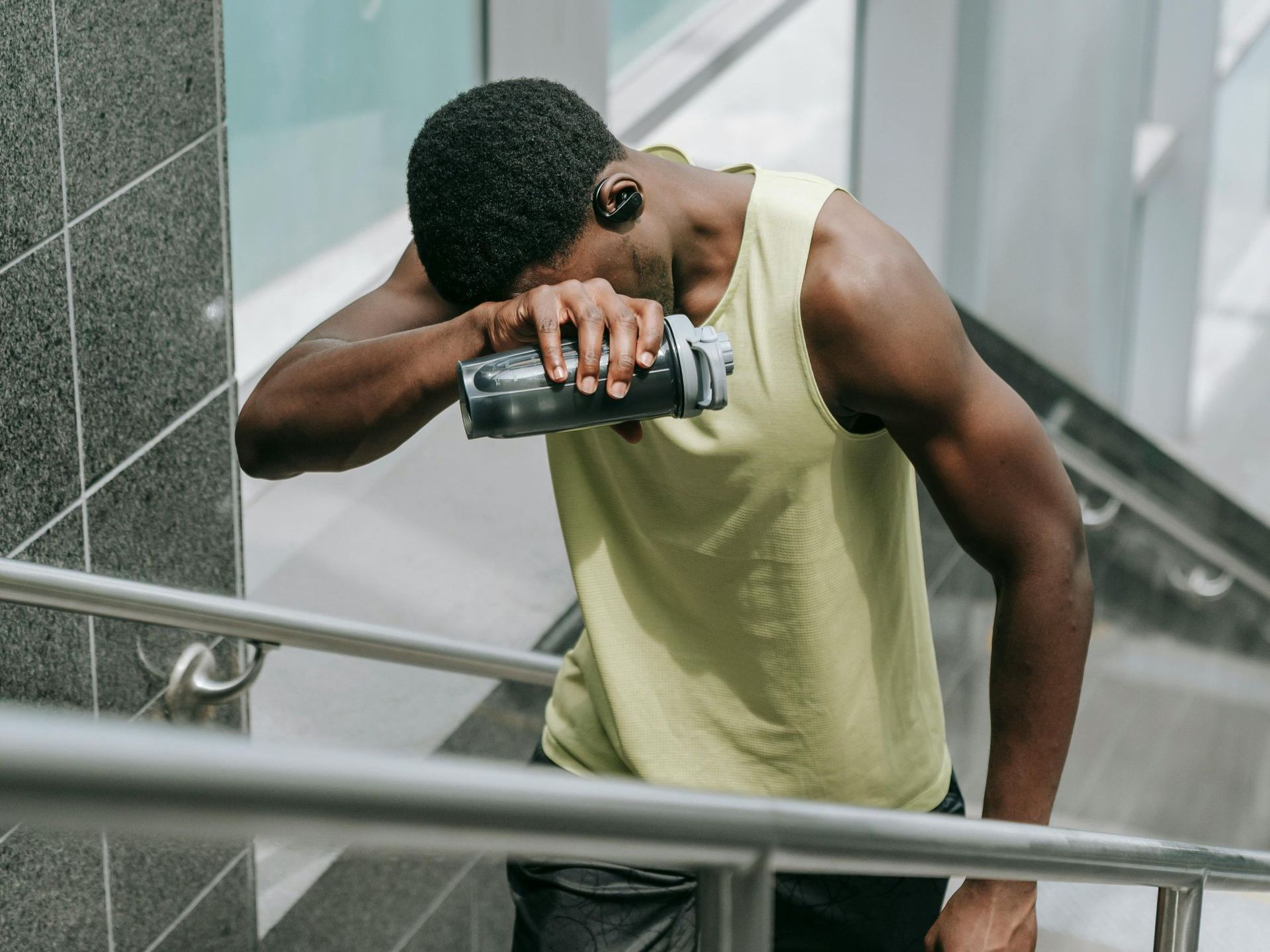 Man in yellow tank top, wiping sweat, pausing on stairs with water bottle.