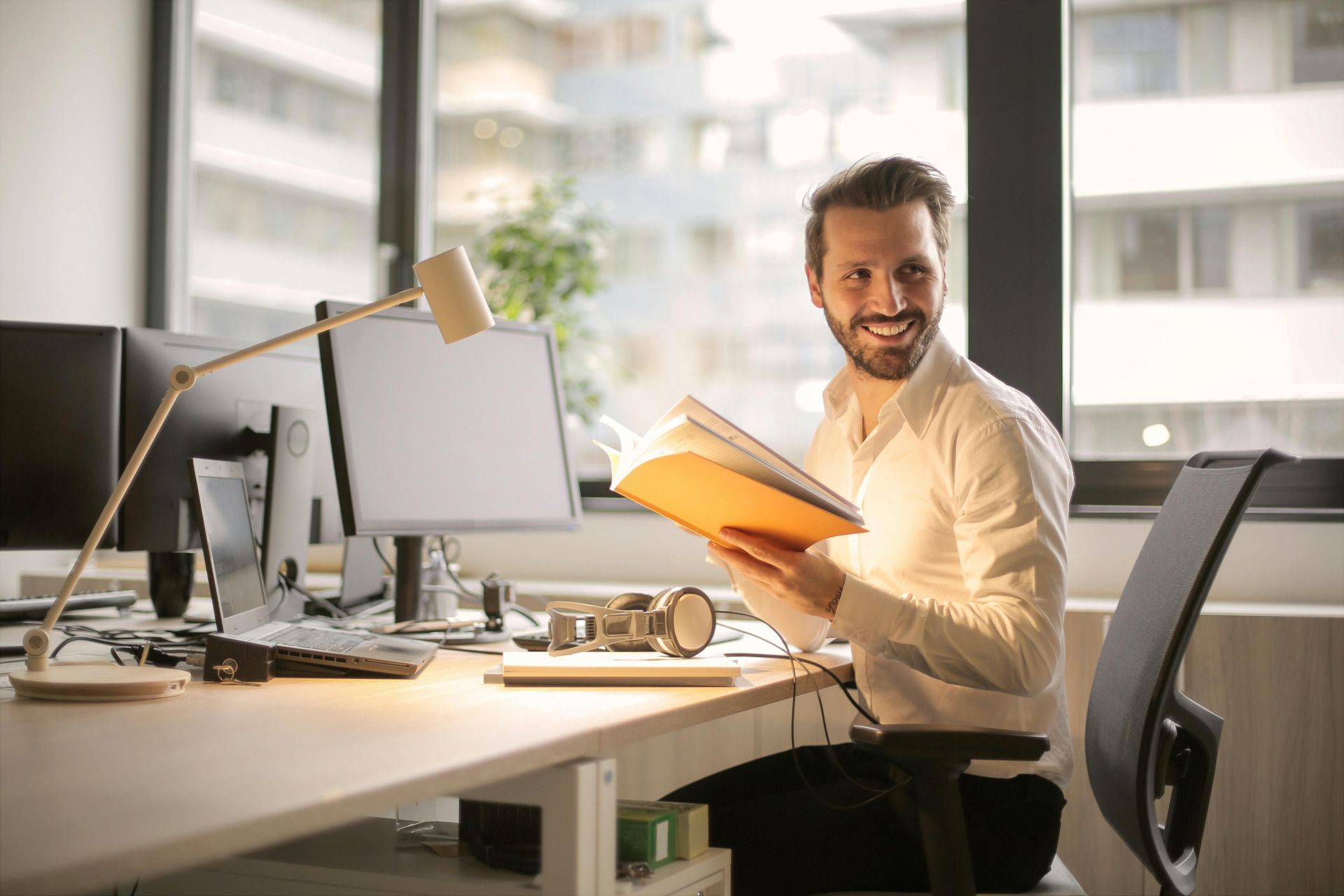 Man smiles while reading a book at his office desk, with a computer and lamp.