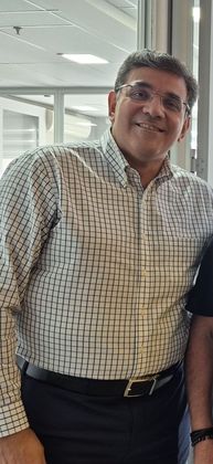 Uma pessoa sorridente usando óculos e uma camisa xadrez clara de botões, por dentro de calças escuras.