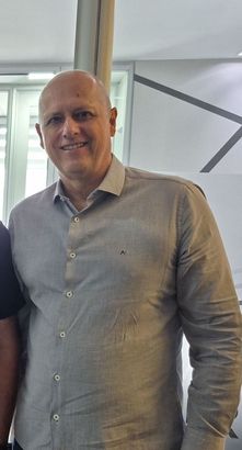 Uma pessoa sorridente, vestindo uma camisa cinza-clara de botões, está em pé em um escritório bem iluminado.