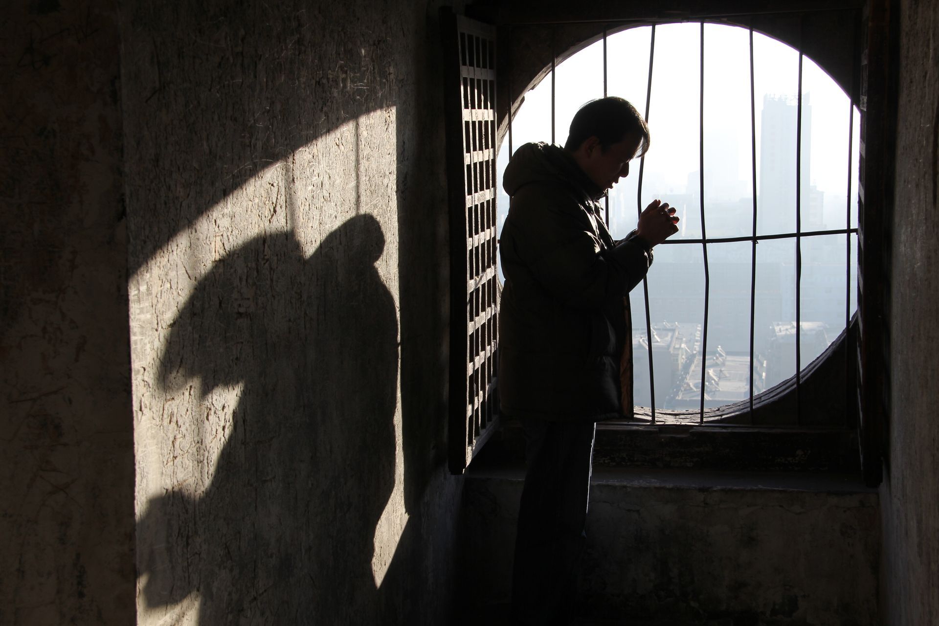 Silhouette of a person praying by barred window, with shadow cast on a wall.