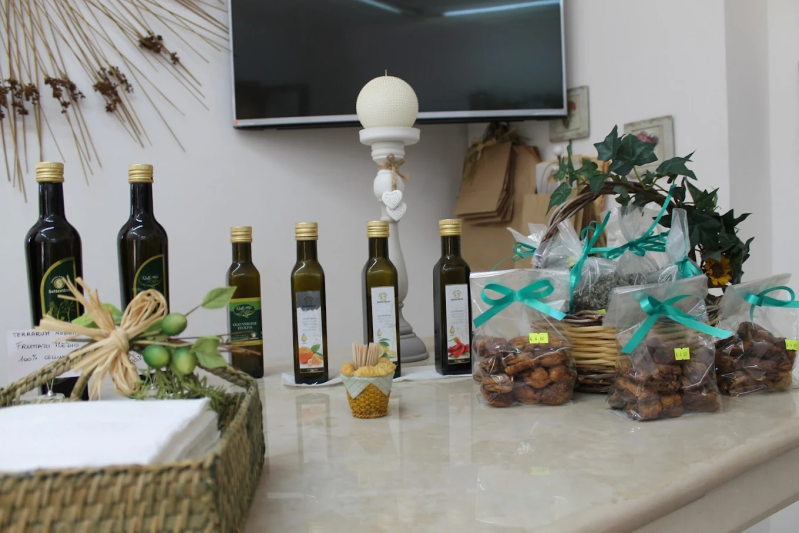 Esposizione di bottiglie di olio d'oliva e dolcetti confezionati su un bancone, con un portacandele decorativo e una piccola TV sopra.