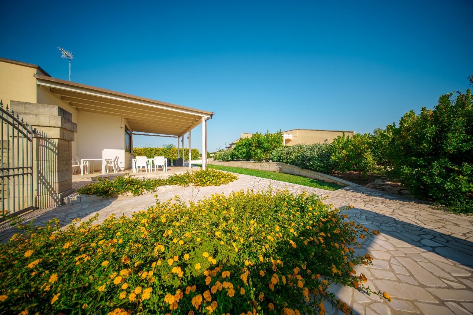 Un bungalow bianco con patio, circondato da fiori gialli e vegetazione lussureggiante, sotto un cielo azzurro.