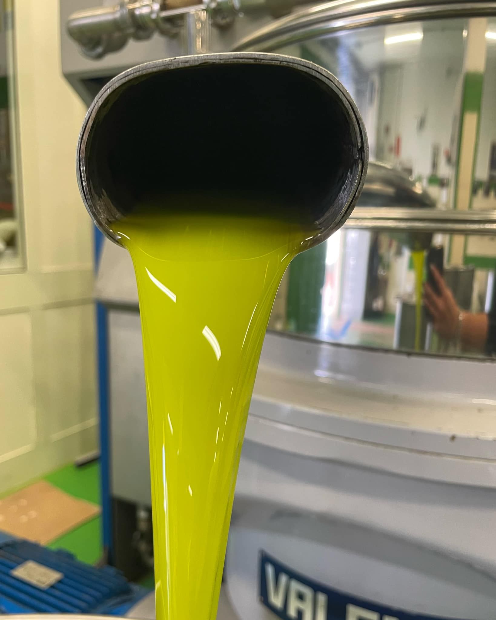 Olio d'oliva che fuoriesce da un beccuccio metallico, di un vivace colore verde-giallo. Ambiente industriale con serbatoio metallico.