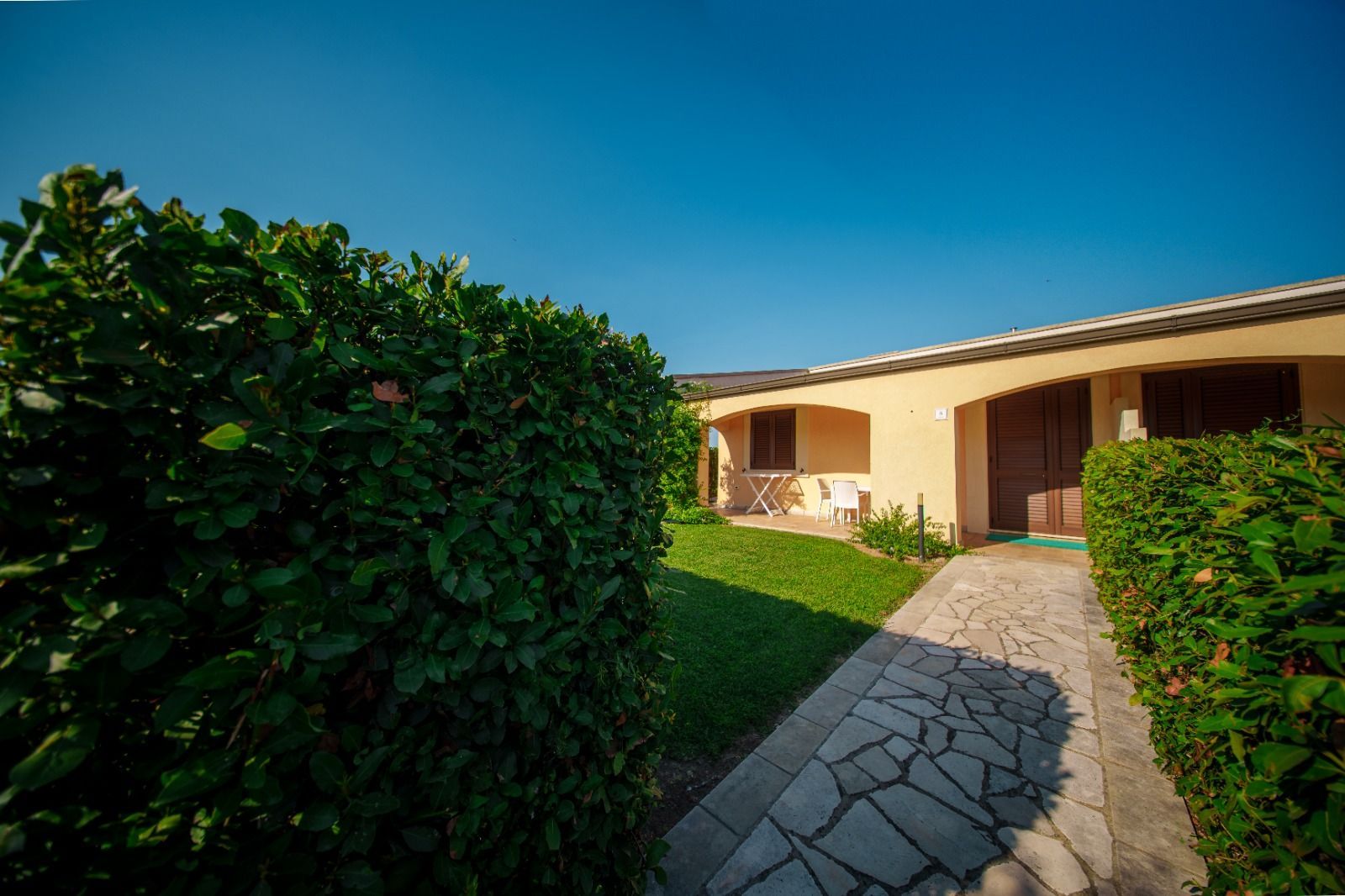 Un sentiero di pietra conduce a edifici beige con porte marroni, fiancheggiati da siepi verdi sotto un cielo azzurro.