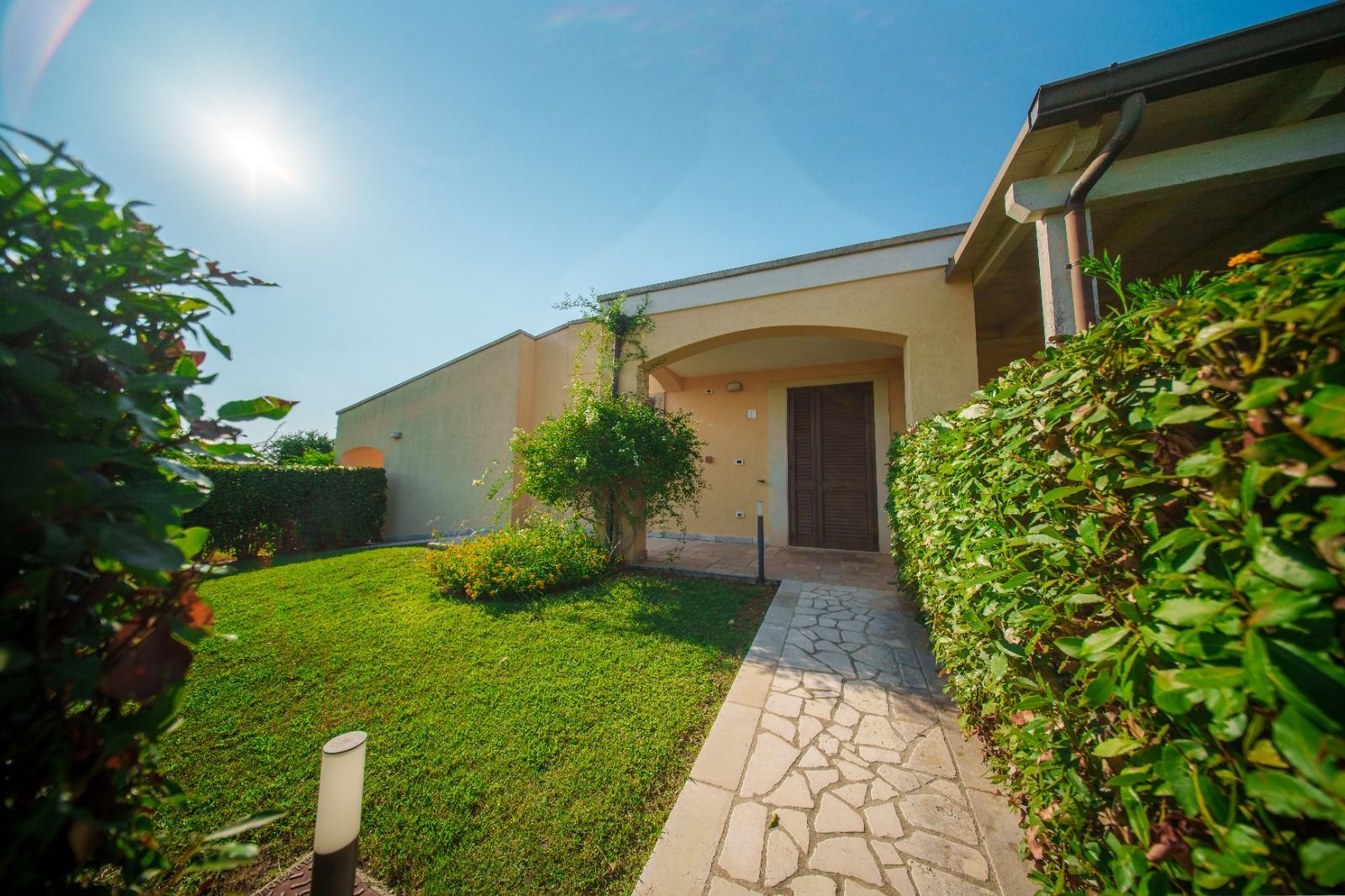 Ingresso di un edificio beige con un sentiero in pietra, fiancheggiato da siepi verdi e un cielo azzurro e soleggiato.