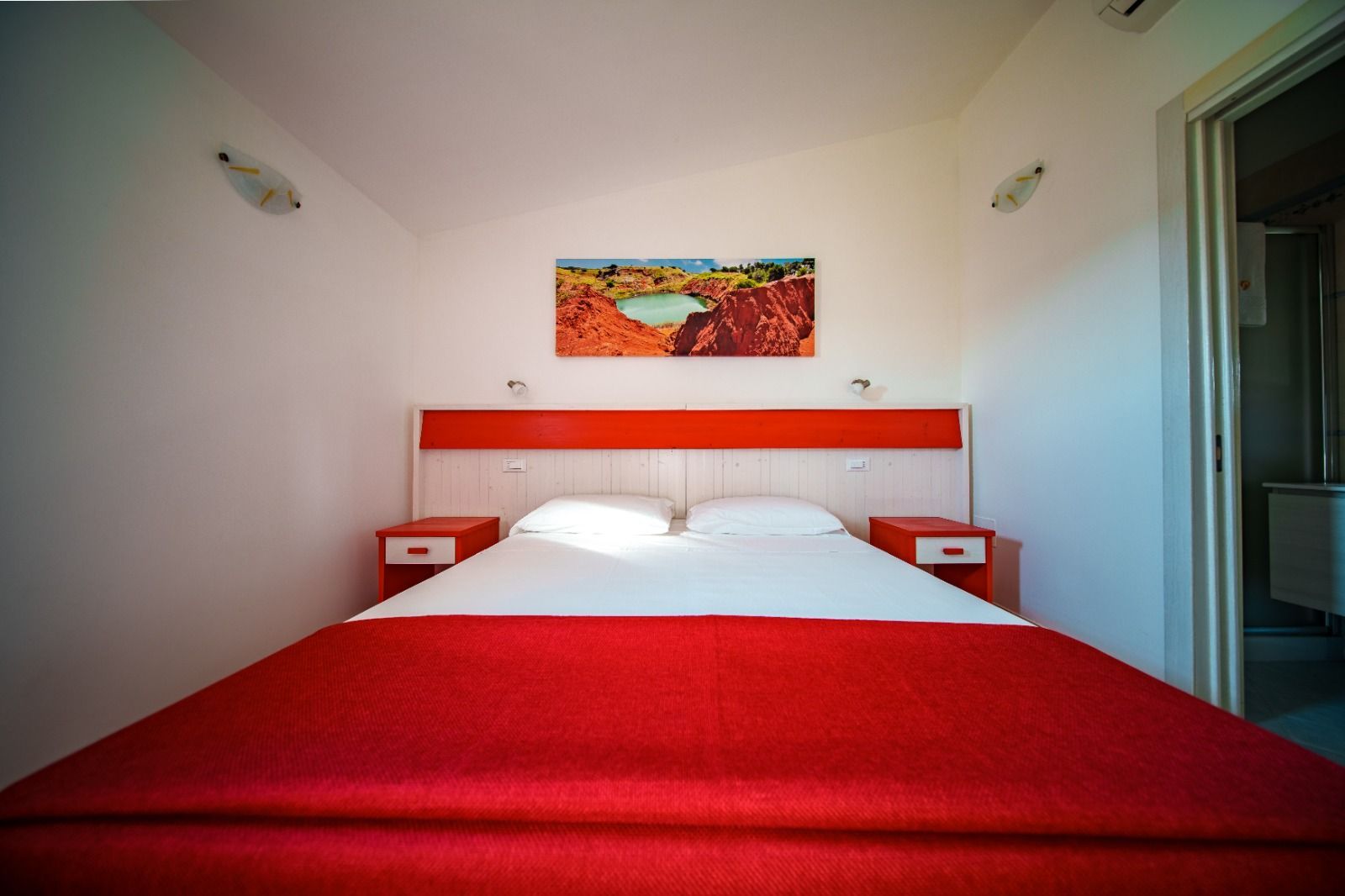 Camera da letto con copriletto rosso, testiera arancione, due comodini bianchi e un dipinto di un paesaggio.