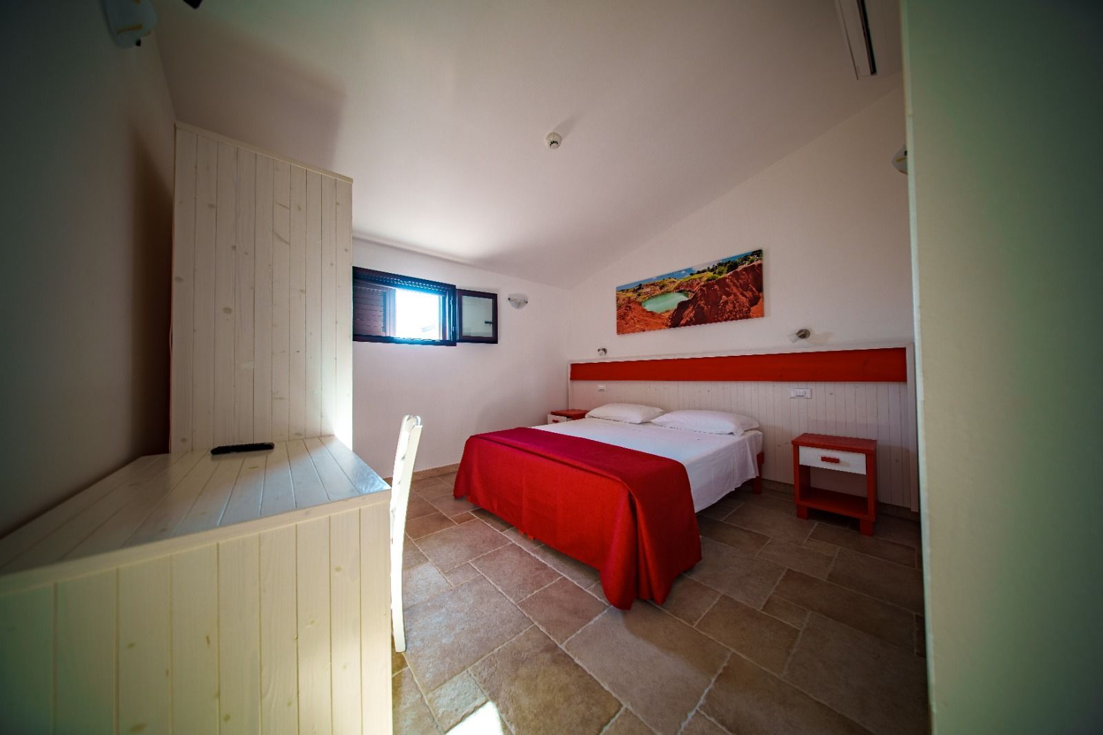 Camera da letto con copriletto rosso, scrivania, piccola finestra, quadri alle pareti e pavimento piastrellato.