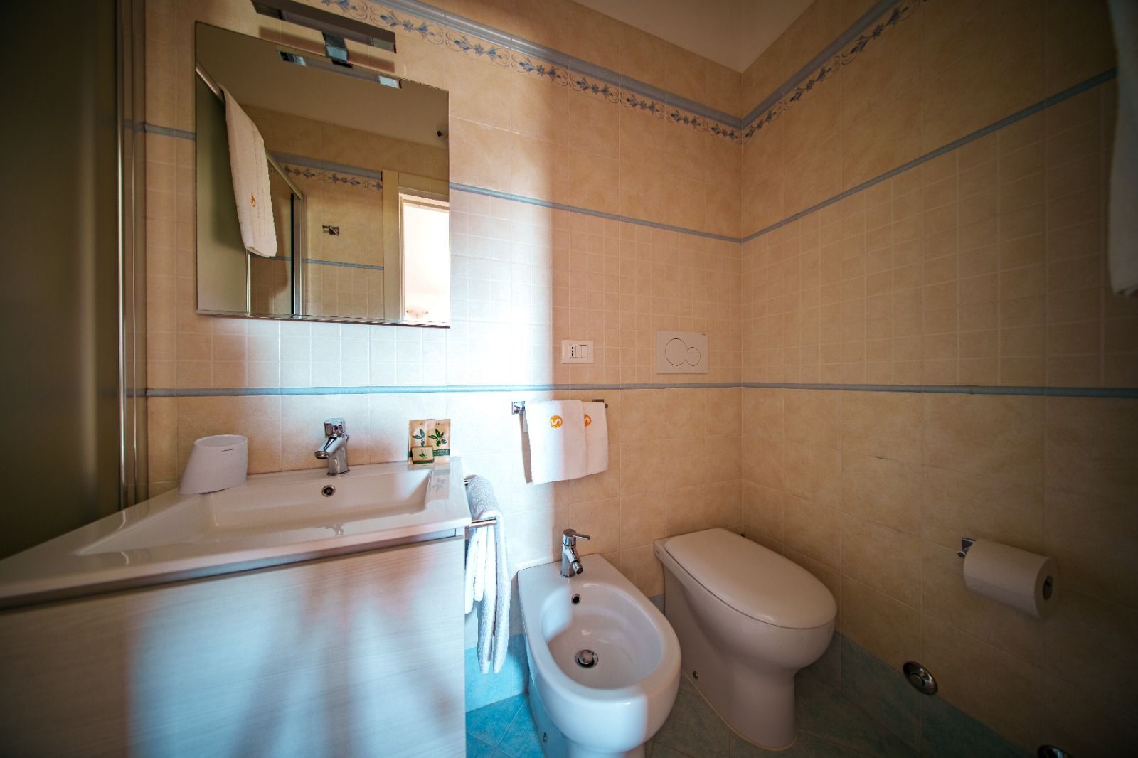 Bagno con lavandino, WC, bidet e specchio. Pareti piastrellate beige e finiture blu.