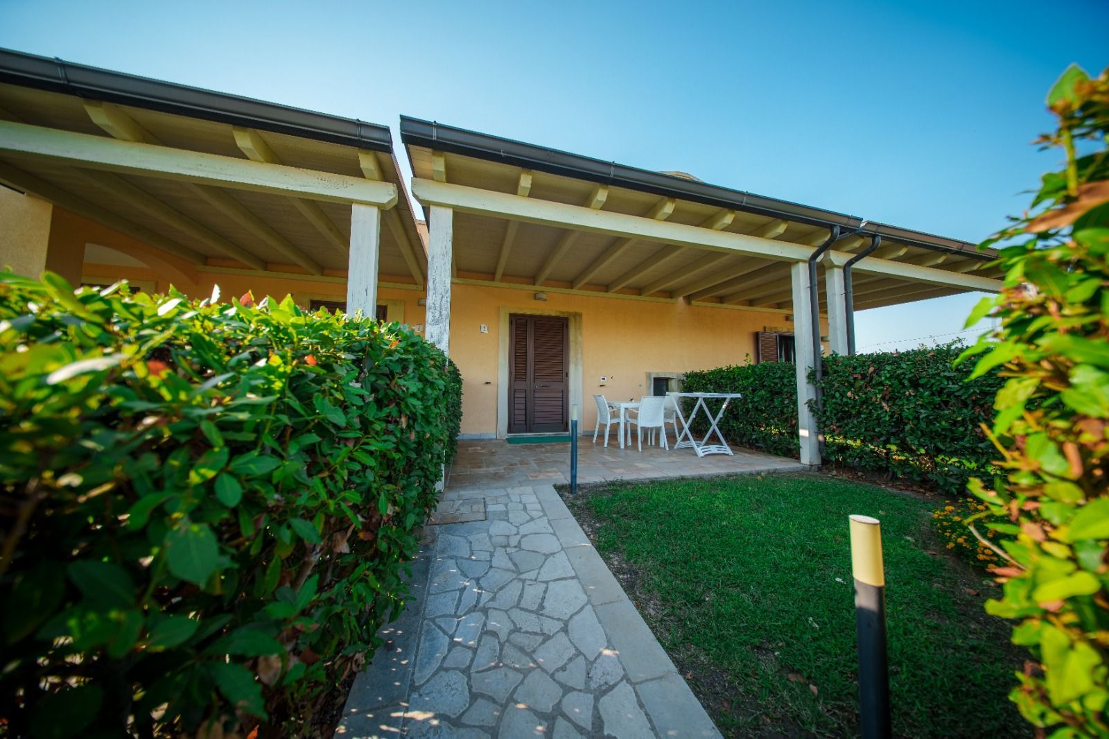 Sentiero in pietra che conduce a una casa con portico, prato verde e arbusti sotto un cielo azzurro.