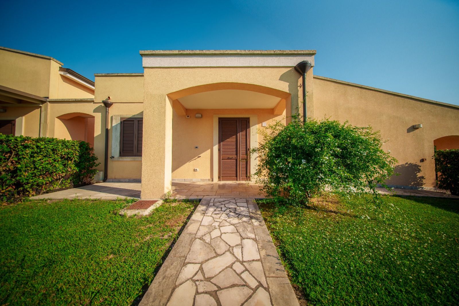 Casa in stucco color marrone chiaro con vialetto in pietra, prato verde, cielo azzurro.