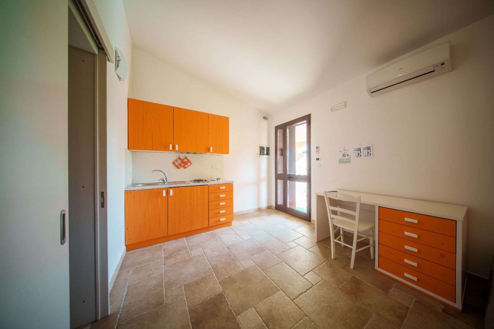 Piccola cucina con mobili arancioni, pavimento in piastrelle di pietra, una scrivania e una porta che dà sull'esterno.