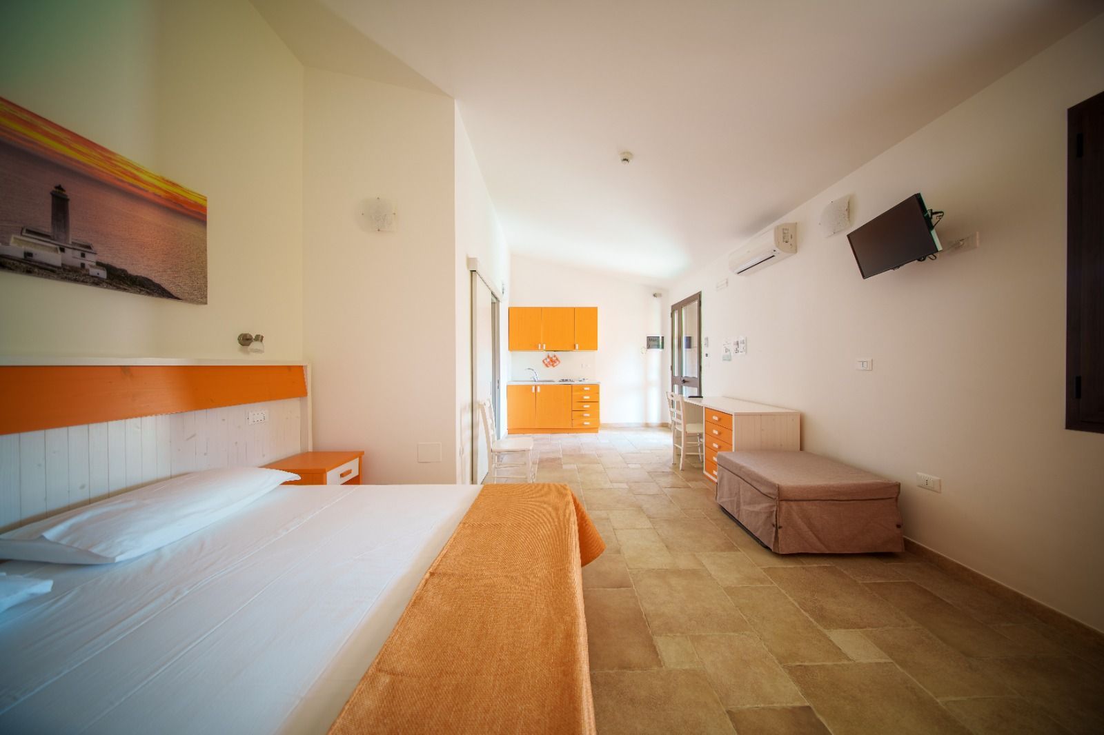 Camera da letto con dettagli arancioni e bianchi, letto, cassettiera, angolo cottura visibile attraverso una porta aperta.