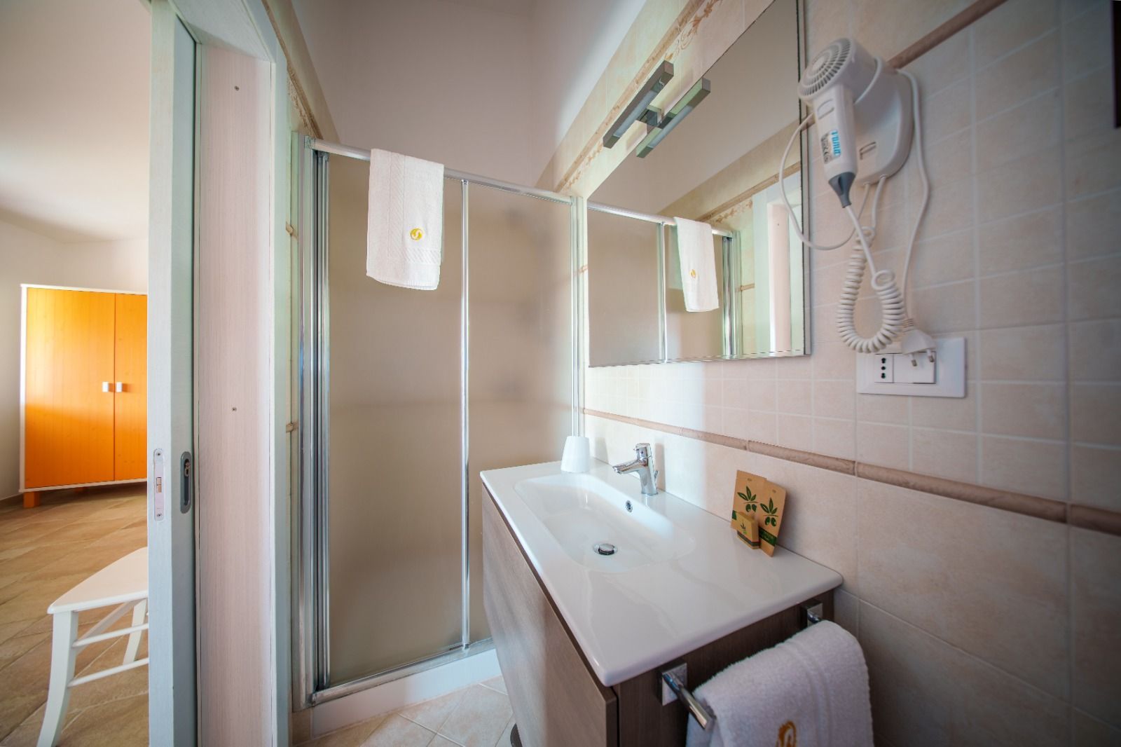 Bagno con lavandino bianco, doccia e specchio. Un asciugamano è appeso al portasciugamani. Una porta aperta conduce alla camera da letto.