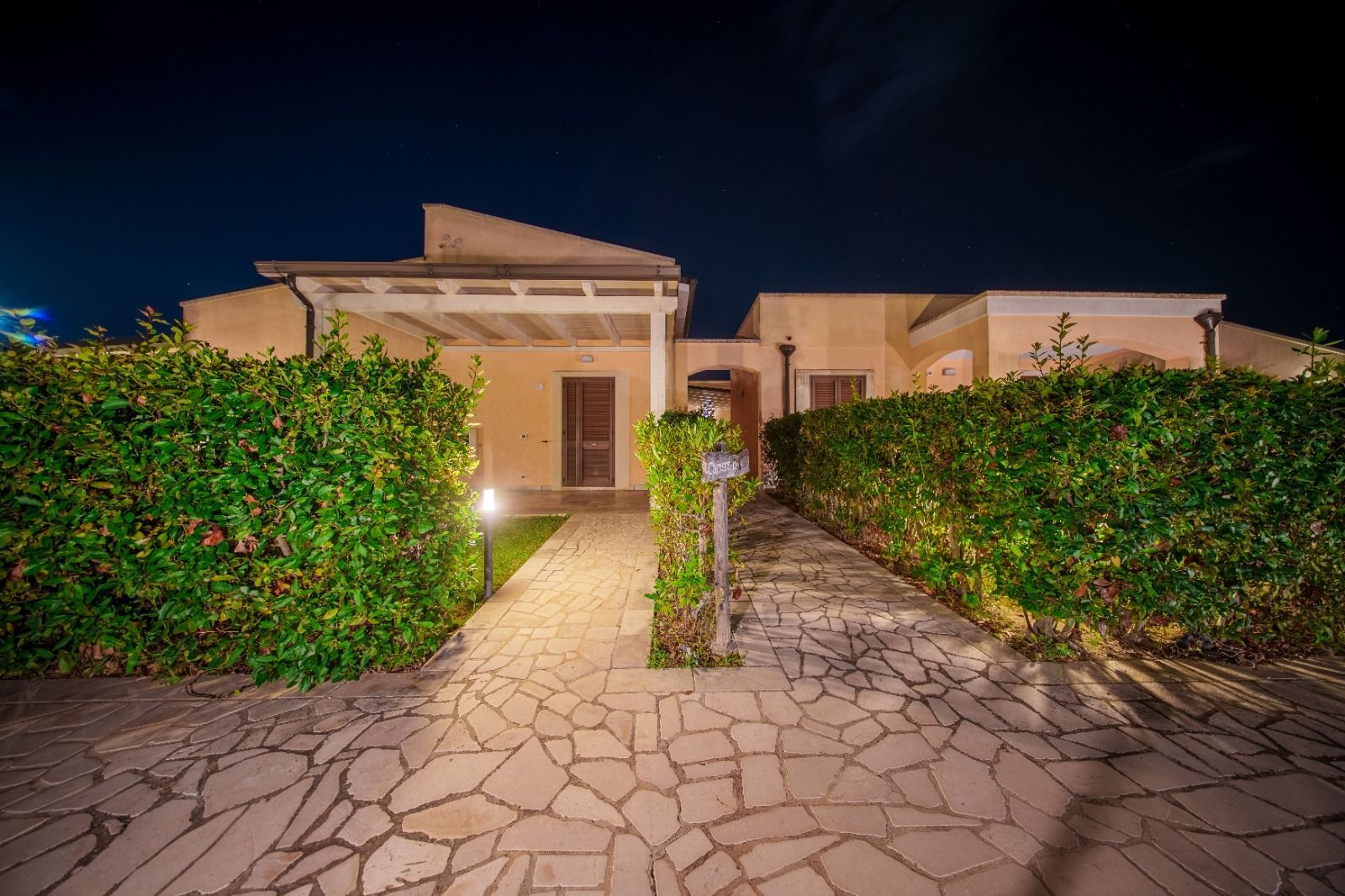 Sentiero in pietra che conduce a un edificio con siepi su entrambi i lati, illuminato di notte da luci esterne.