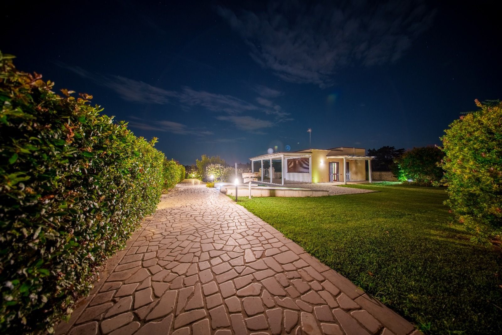 Vista notturna di un sentiero di mattoni che conduce a una casa ben illuminata con un pergolato bianco, fiancheggiato da cespugli ed erba verde sotto un cielo scuro.