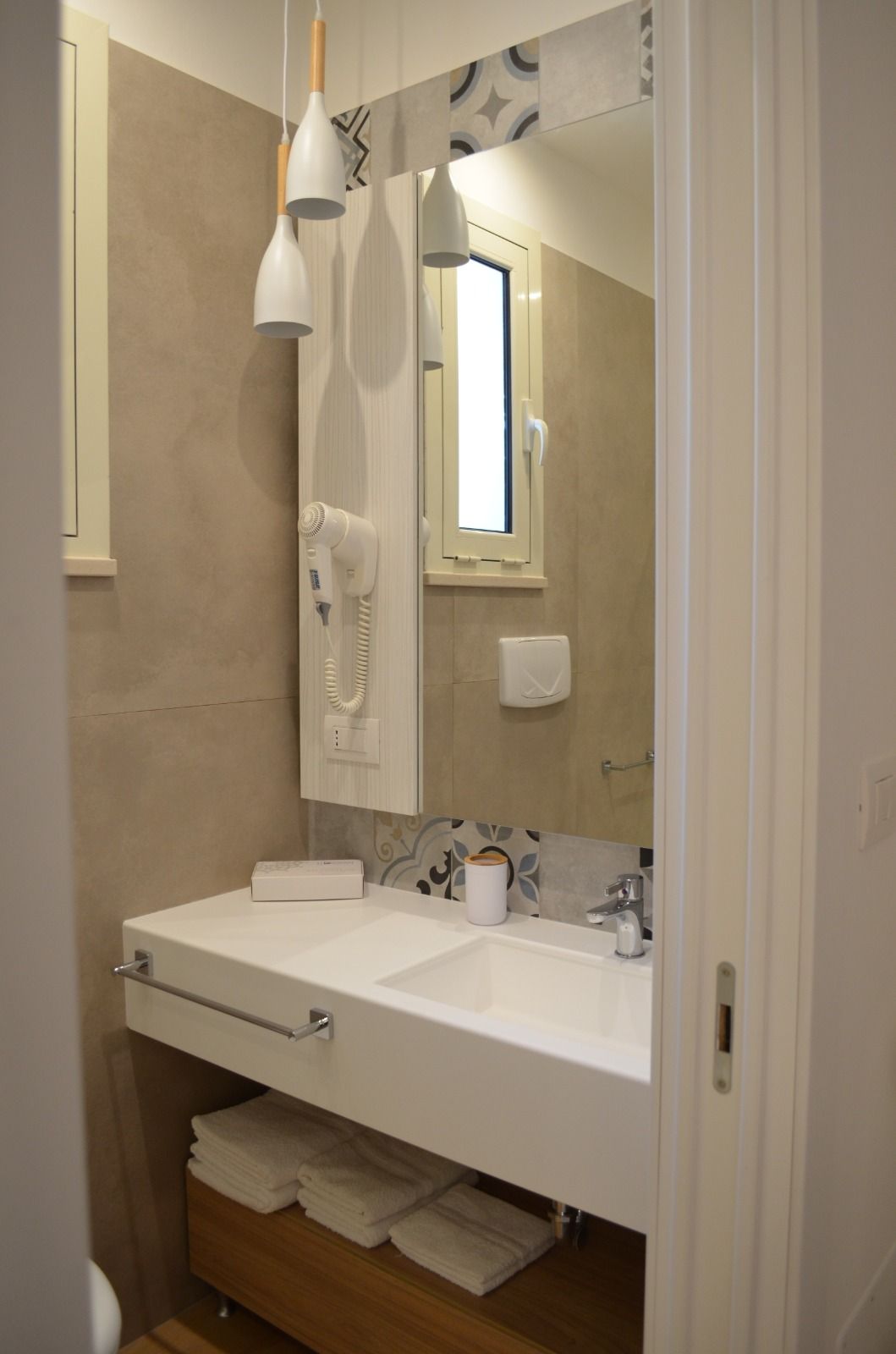 Bagno con lavandino bianco, specchio e una piccola finestra. Pareti beige e lampade a sospensione bianche.