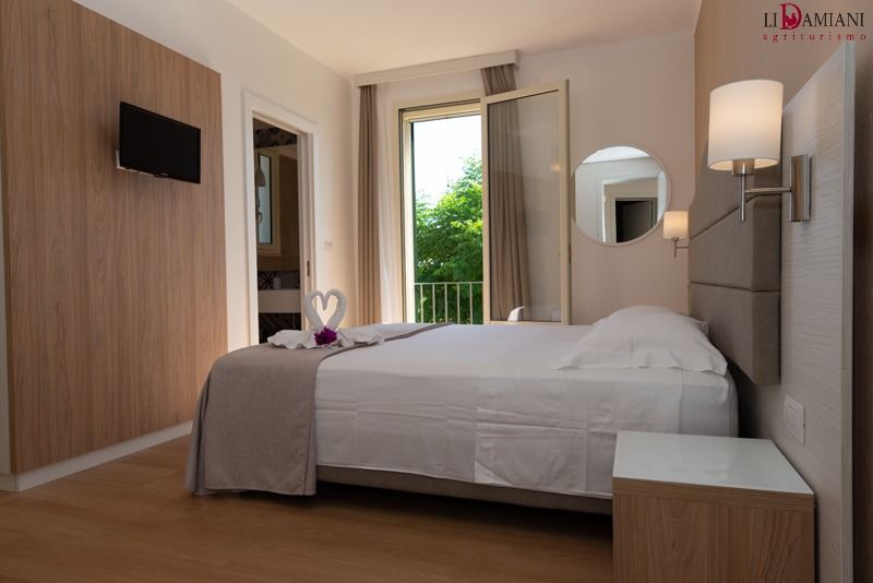 Camera d'albergo con letto, TV e balcone. Pannelli in legno chiaro, lenzuola bianche e arredamento dai toni neutri.