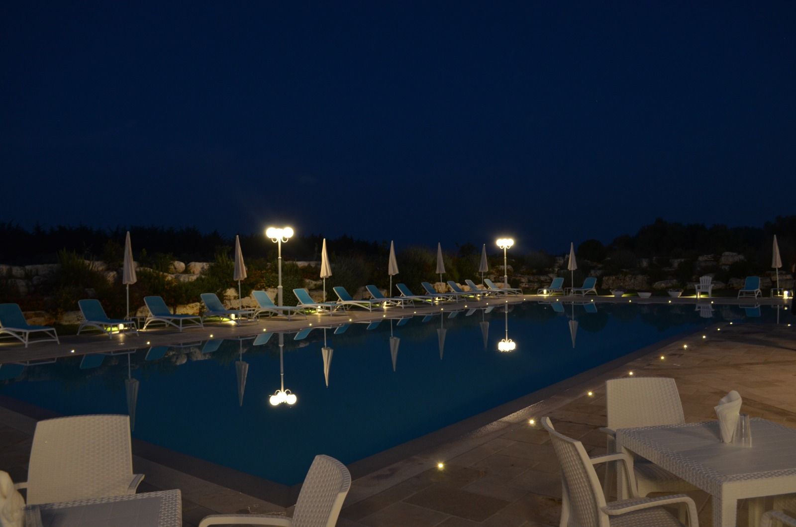 Una piscina di notte, illuminata da lampade a soffitto e luci a bordo piscina, con lettini e tavolini in primo piano.