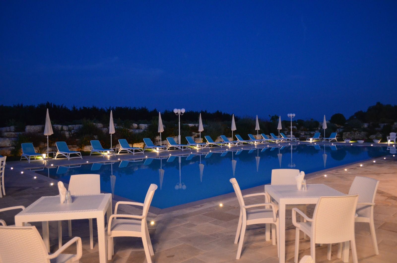 Posti a sedere a bordo piscina al tramonto, tavoli e sedie bianchi, piscina illuminata, sedie a sdraio, cielo blu intenso.