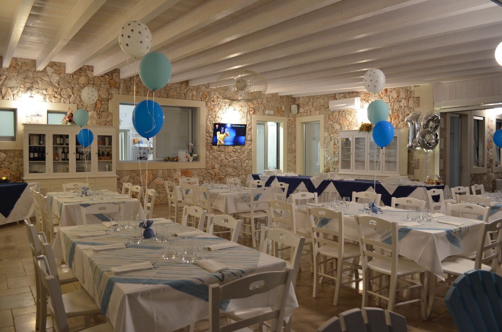 Sala da pranzo allestita per una festa con palloncini blu e bianchi, tavoli con runner blu e sedie di legno bianche.