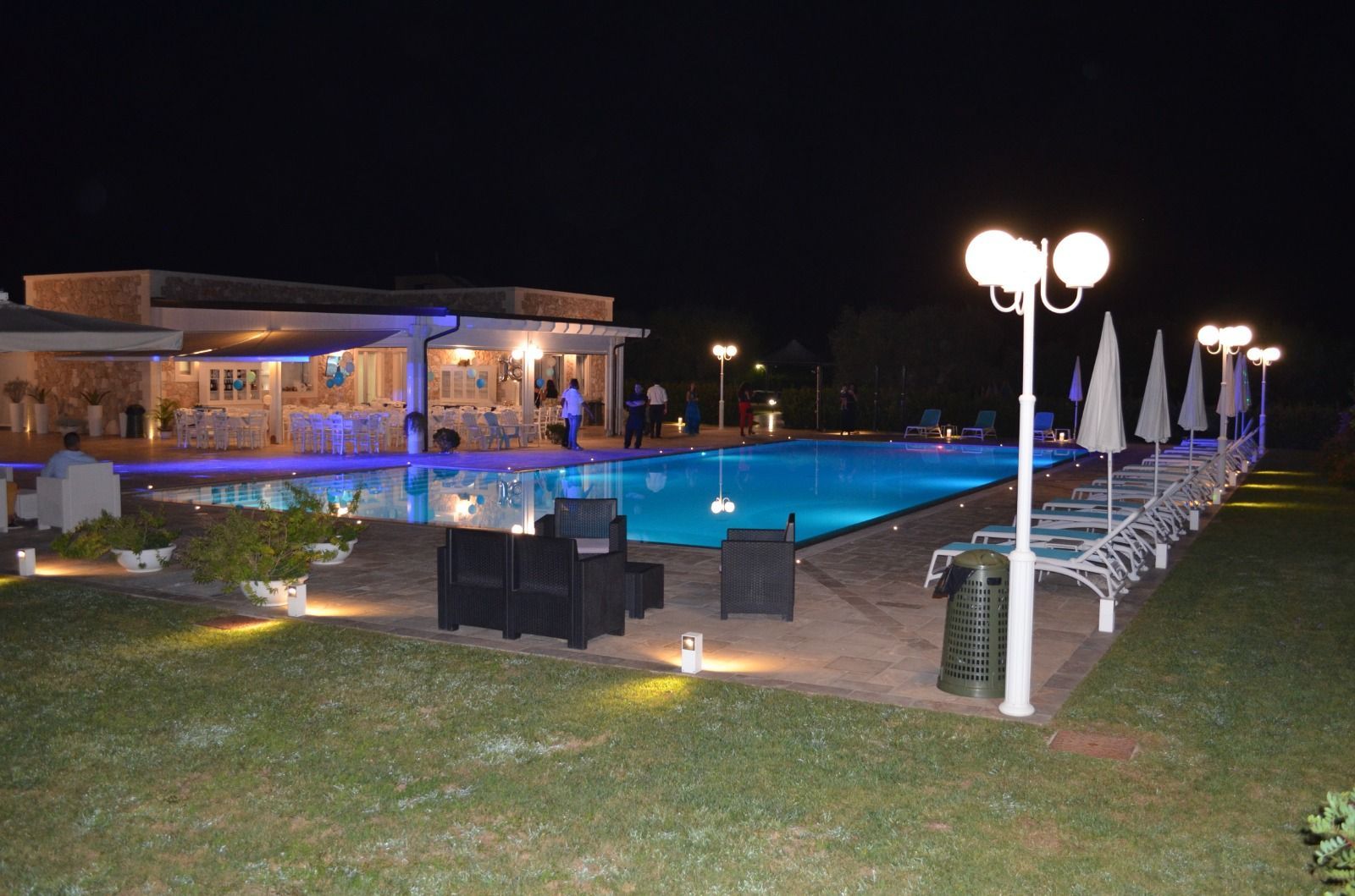 Vista notturna di una piscina con area circostante illuminata, edificio e lampioni bianchi.