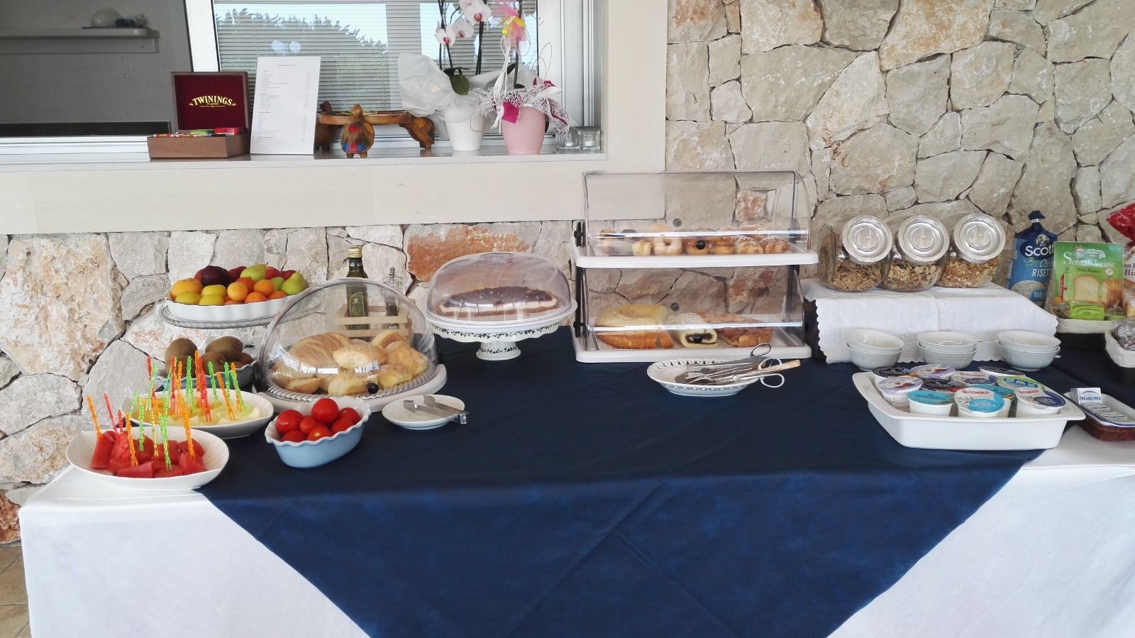 Un tavolo imbandito con pasticcini, frutta e snack per la colazione, sullo sfondo di un muro di pietra.