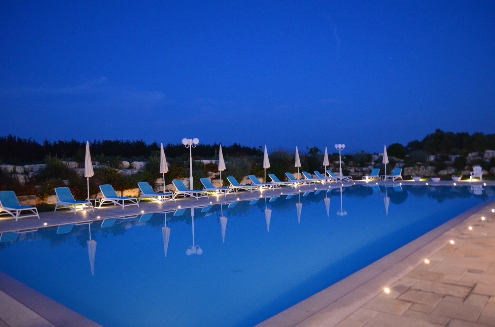 Piscina al tramonto con sdraio e ombrelloni blu che si riflettono nell'acqua.