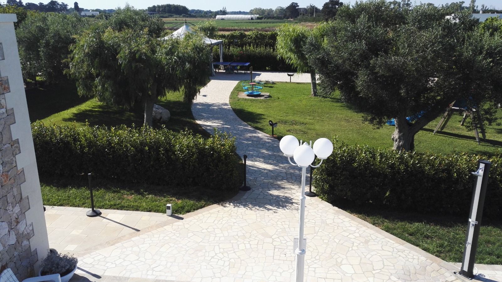 Sentiero in pietra attraverso un prato verde con alberi, che conduce a una struttura in lontananza. Palloncini bianchi su un palo in primo piano.