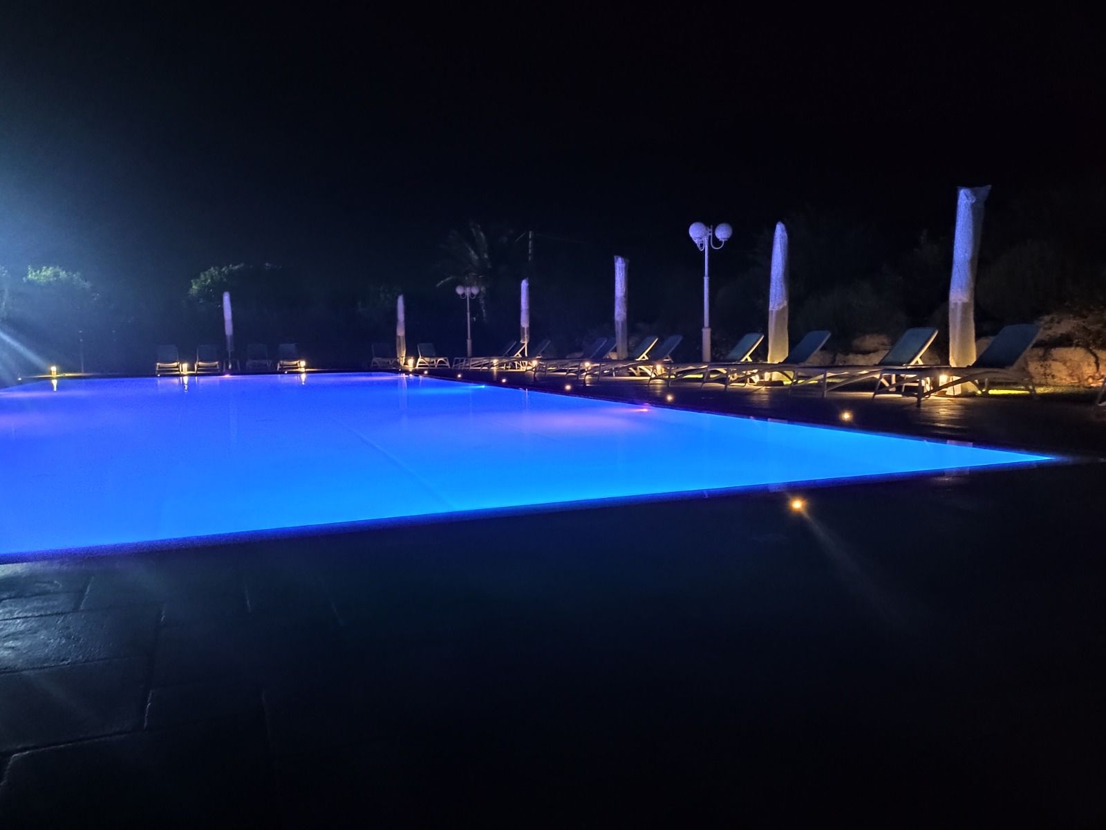 Ripresa notturna di una piscina a sfioro illuminata di blu, sedie a sdraio e colonne decorative.