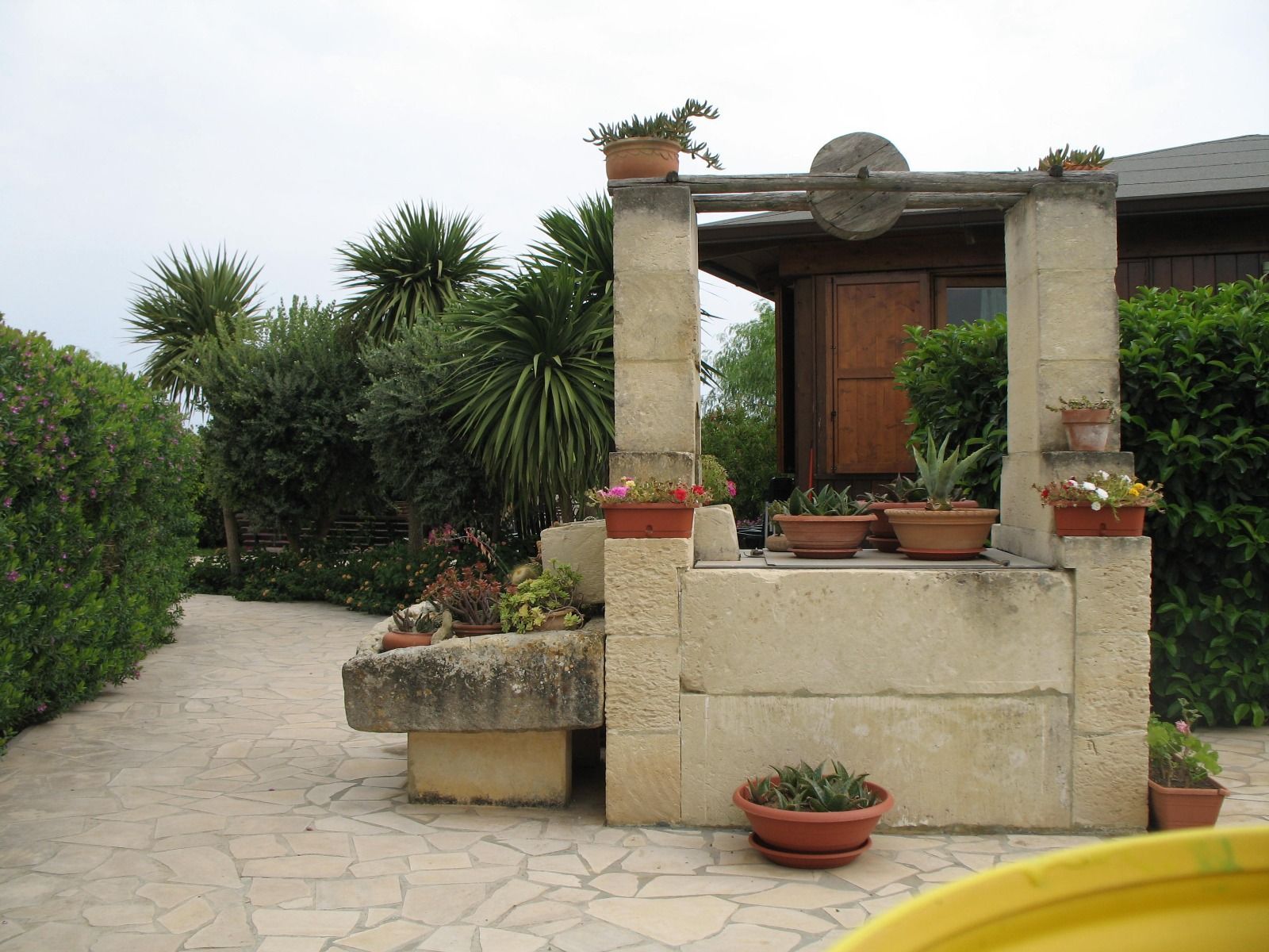 Pozzo in pietra con piante in vaso, circondato dal verde e da un sentiero lastricato. Cielo coperto.