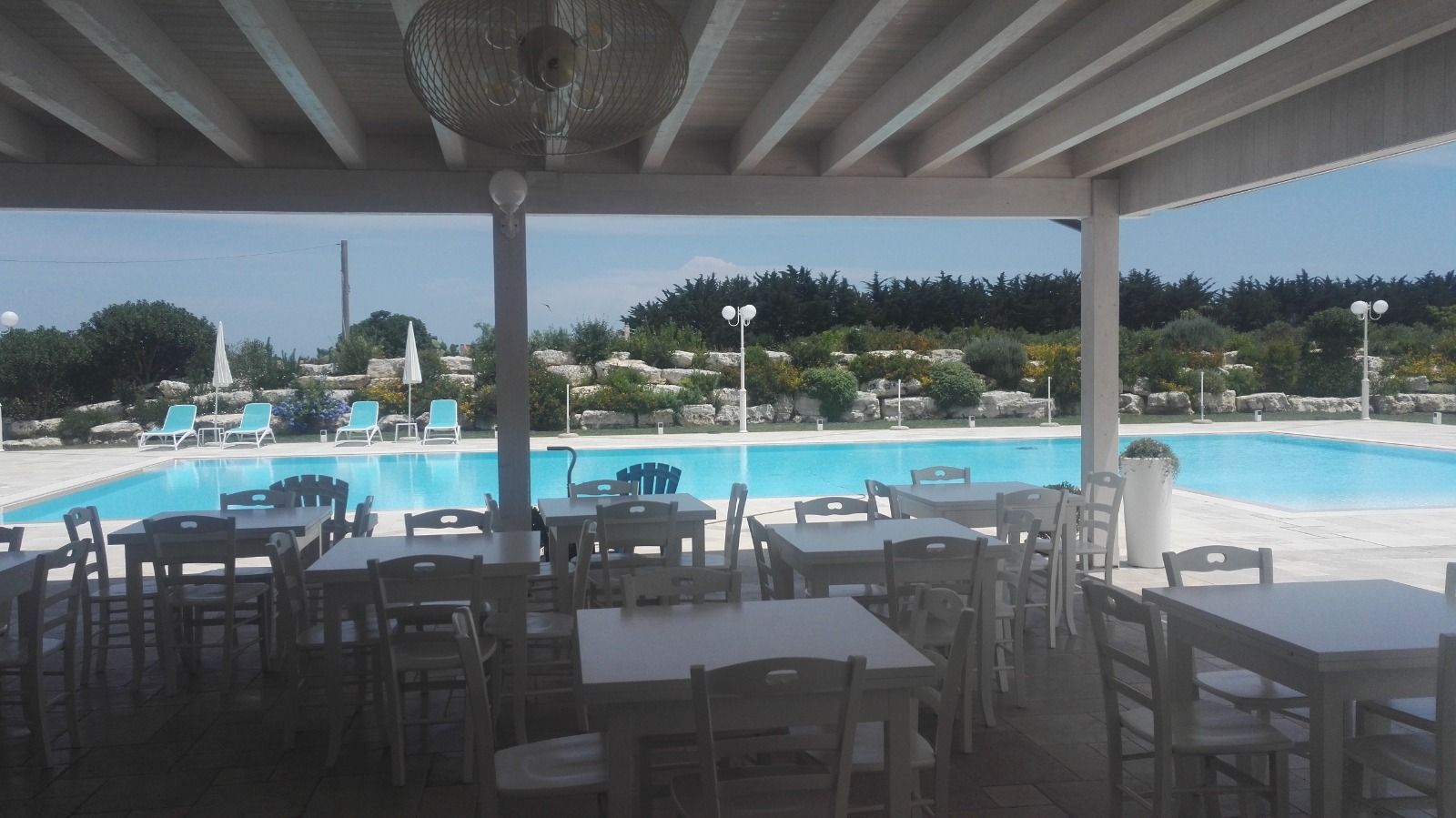 Tavoli e sedie su una terrazza bianca si affacciano su una grande piscina blu e sul verde circostante.