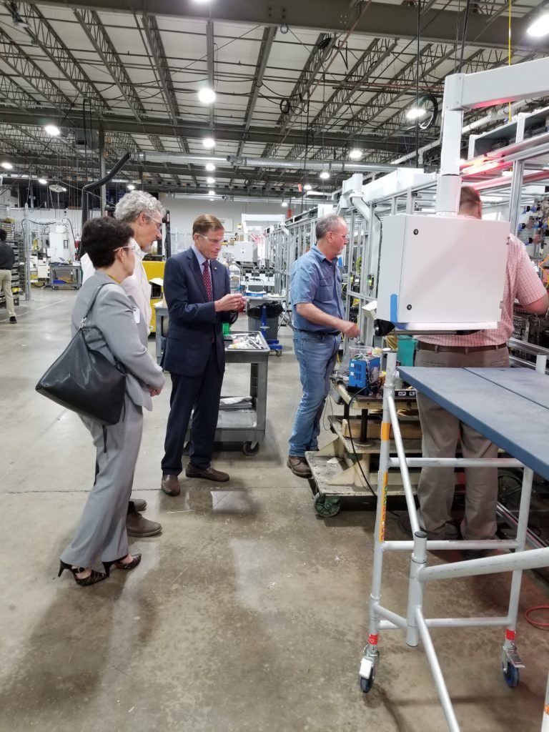 People in a factory setting observe machinery and a worker operating it.