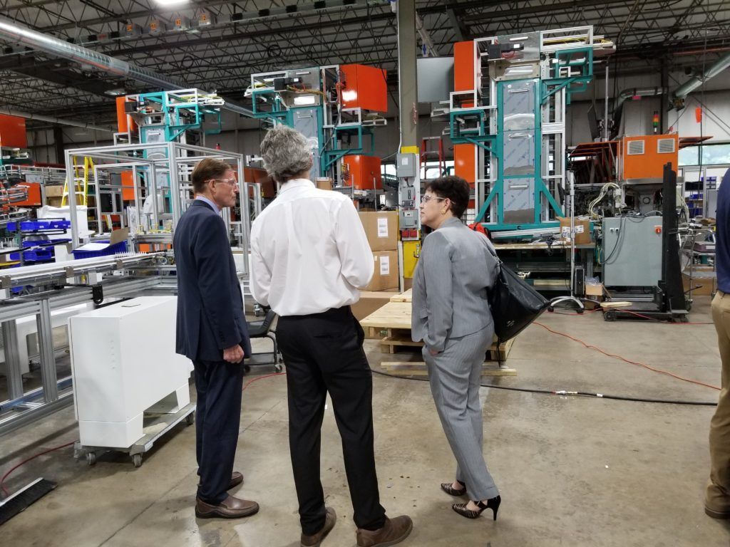 Three people in suits talking near industrial machinery in a factory setting.