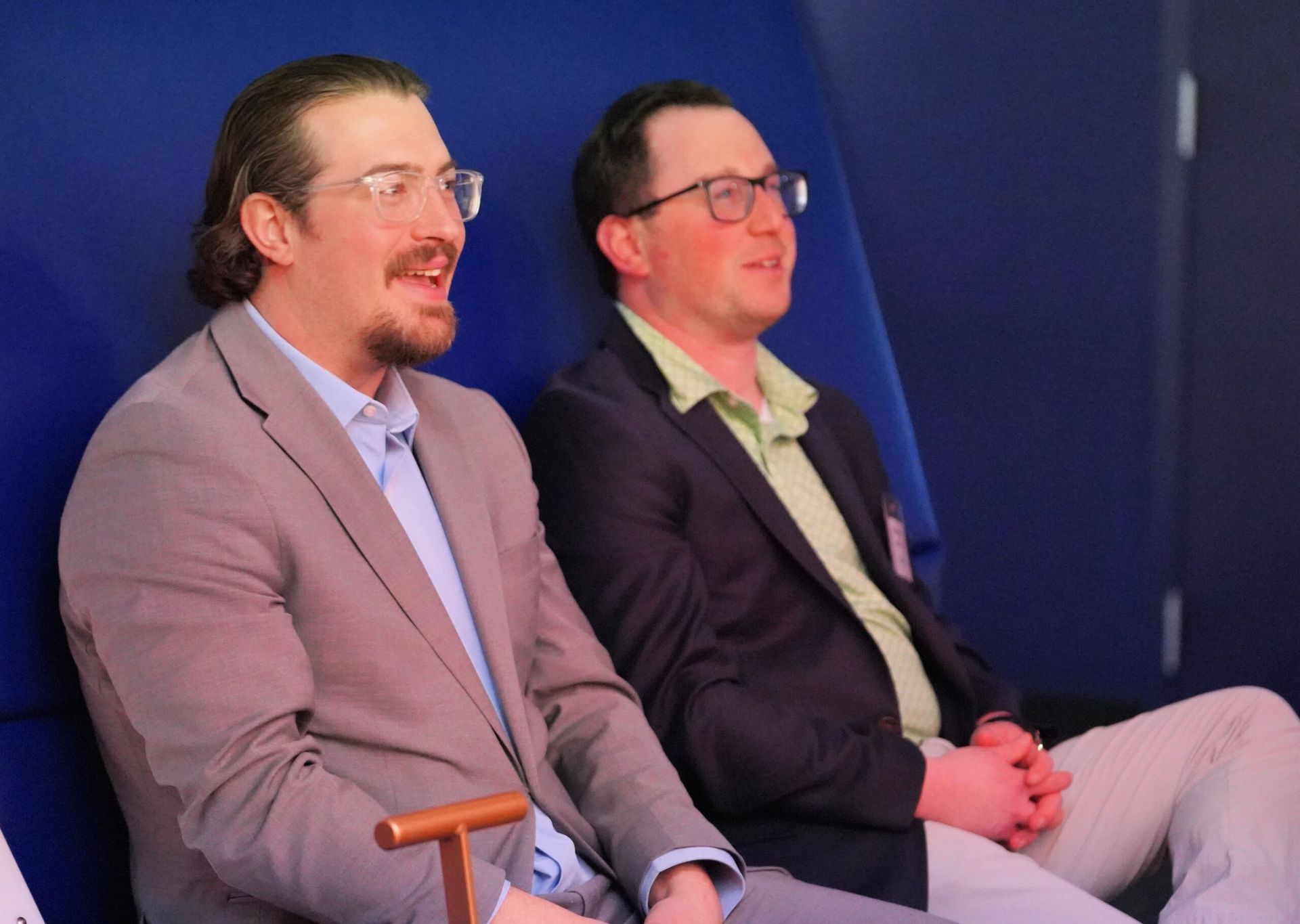 Two men in suits seated, one speaking, the other listening in a blue setting.