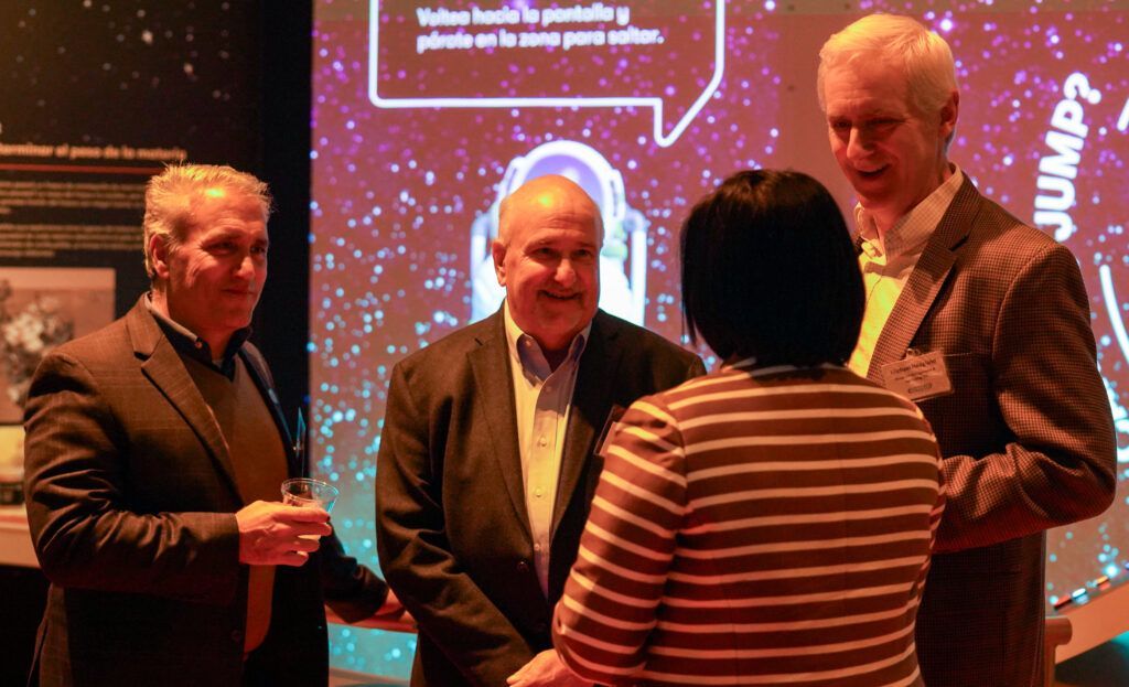 Four people converse near a display screen at an event. The screen has text in a foreign language and the word 