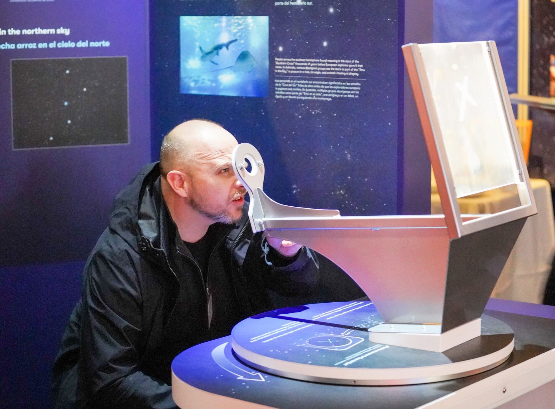 Man looking through a silver exhibit, possibly a telescope. Dark-haired, in a black jacket. Blue display in background.